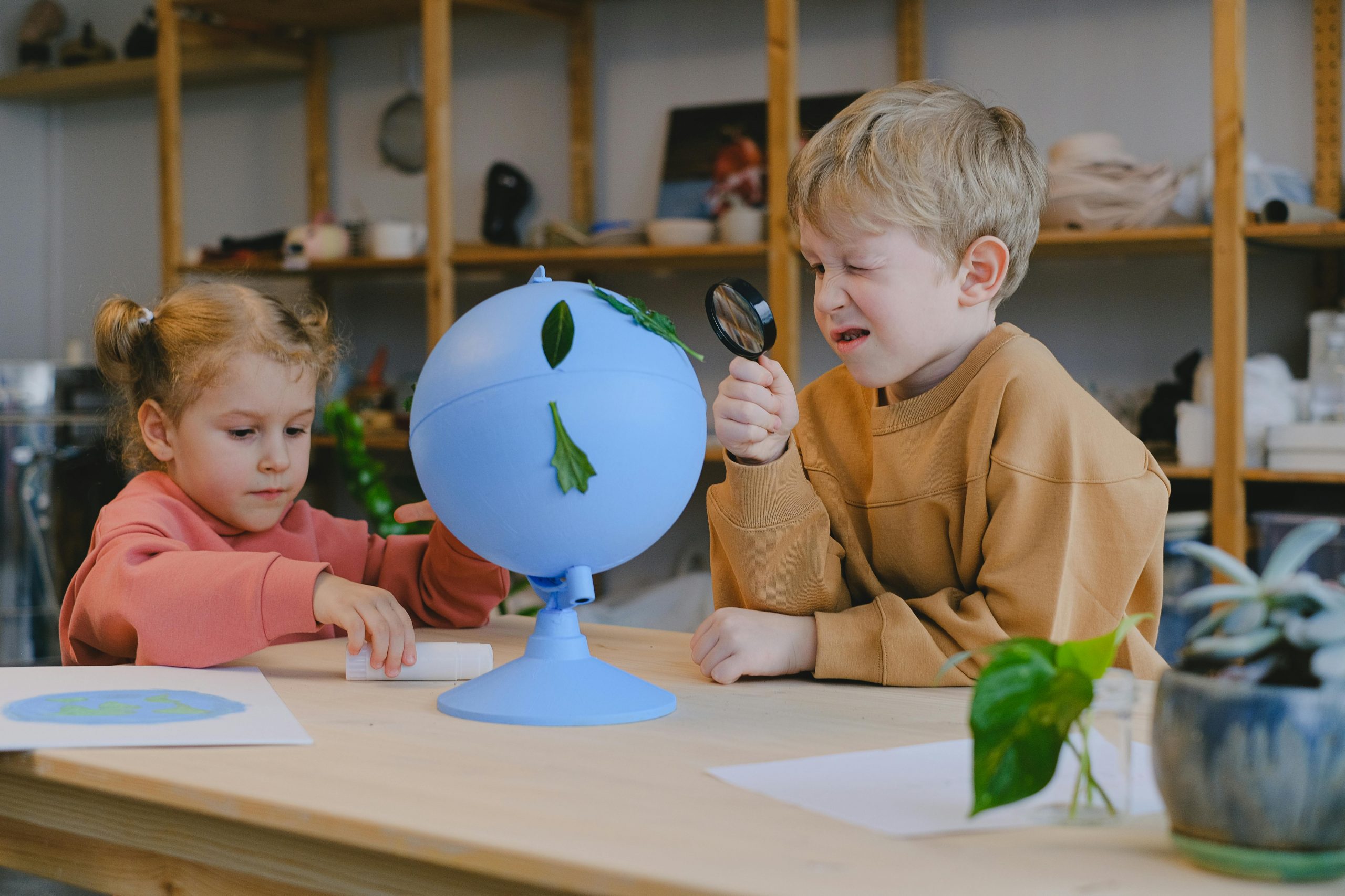 Two young children exploring a blue globe with leaves using a magnifying glass during a provocation.
