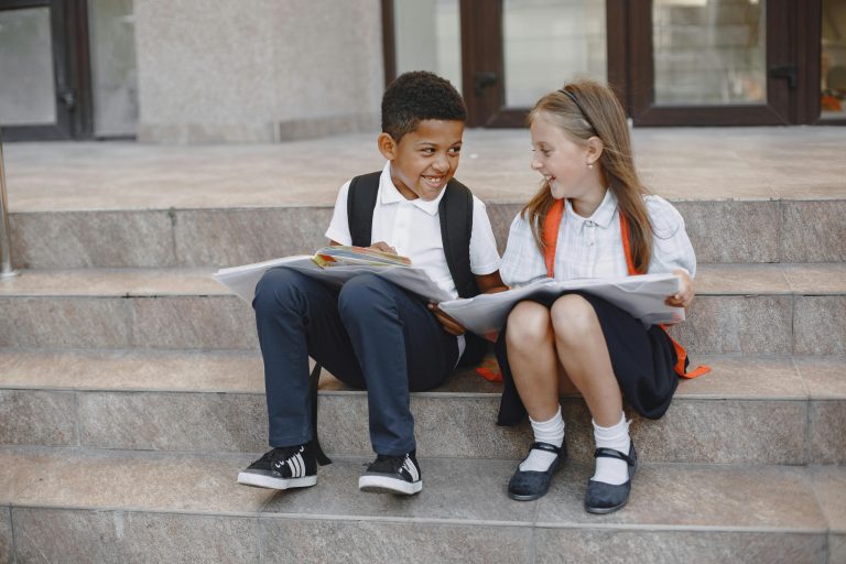 Boy and girl with book bags sitting on school steps, smiling and sharing notebooks — a perfect example of the buddy system for welcoming new students.